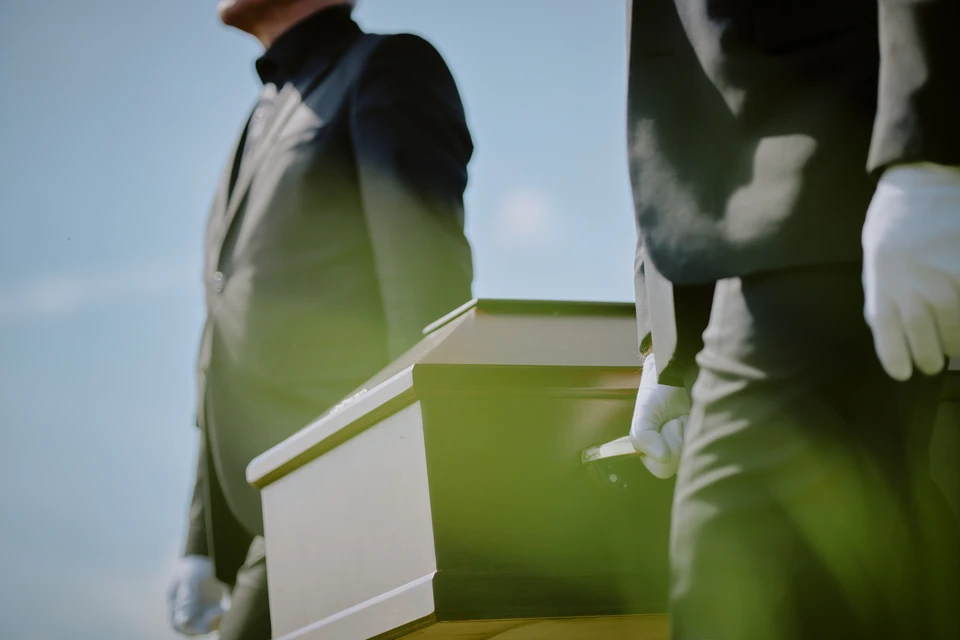 Photographie d'une personne en deuil (femme ou homme d'âge adulte) assise à une table, le visage pensif et légèrement abattu, entourée de documents administratifs et de formulaires étalés devant elle, symbolisant la charge des démarches administratives après un décès. L'ambiance doit être douce et respectueuse, transmettant à la fois le poids des formalités et un sentiment de soutien bienveillant. Éviter tout excès dramatique ou dépressif.